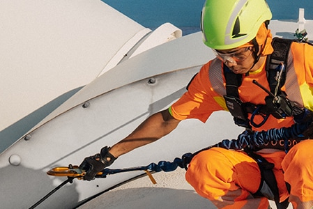 Người đang ngồi chồm hổm trên đỉnh tua-bin gió, trang bị đầy đủ thiết bị bảo vệ chống rơi ngã.