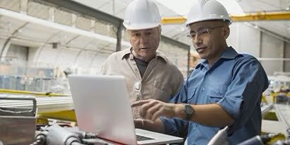 Hình ảnh hai người đàn ông đội mũ cứng vừa thảo luận vừa nhìn vào màn hình laptop
