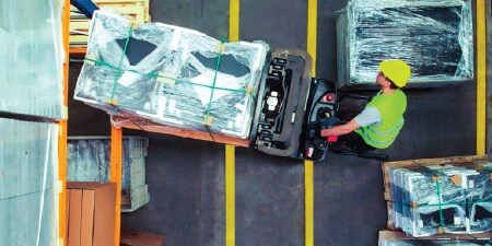 Góc nhìn từ trên xuống về một người điều khiển xe nâng đang di chuyển pallet qua các vạch kẻ bằng băng keo vào khu vực kho hàng
