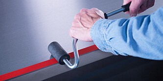 Một công nhân sử dụng một con lăn hình chữ J để ép lên một miếng băng keo trên một thanh kim loại

