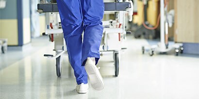 chuyên gia y tế đẩy bệnh nhân trên xe lăn trong hành lang bệnh viện, tiêu điểm nằm ở sàn bệnh viện không có các mối nguy hiểm và được vệ sinh bằng sản phẩm chăm sóc sàn 3M.
