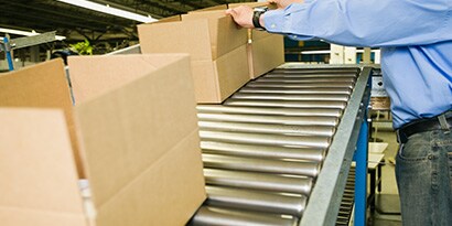 mage of open boxes moving along a conveyor belt to a worker who folds flaps to prepare for sealing
