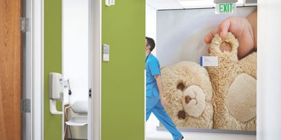 Một nhân viên y tế sải bước qua một bức tường dán lớn, bắt mắt vẽ hình một con gấu bông được bàn tay em bé ôm lấy.

