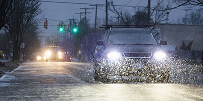A car’s headlights illuminate falling snow on a road at dusk.
